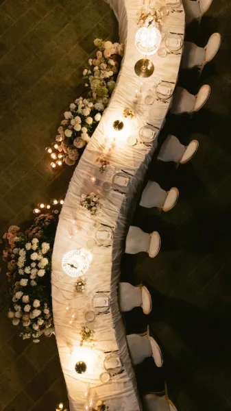 Reception tablescape with long banquet table set in white linens, low florals, candles, glassware, and hanging pendant lights above