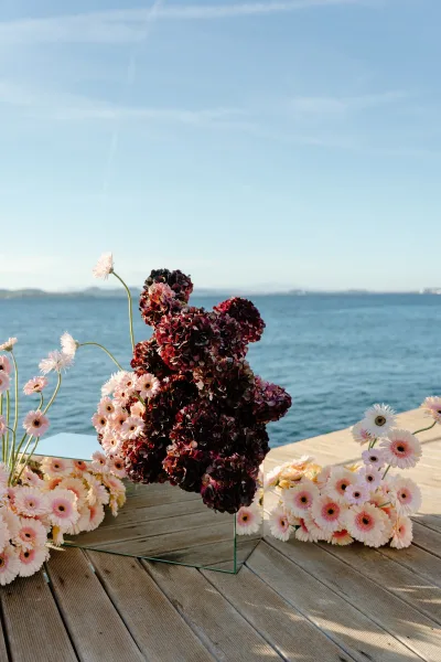 Wedding floral installation with ceremony ground flowers and hydrangeas on a wooden deck beside ocean water, blue sky and distant hills beyond.