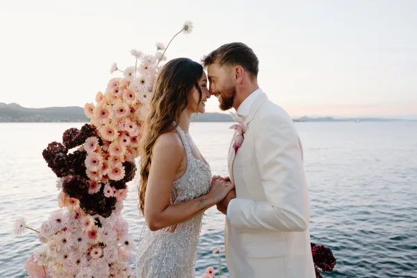 Couple portrait of a bride and groom holding hands beneath a floral arch by the water, sky glowing over distant hills as they touch foreheads.