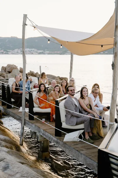 Ceremony guests settle into dockside ceremony seating on lounge sofas along a wooden pier, facing ocean water and a rocky shoreline.