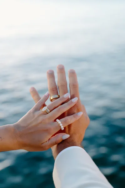 Wedding rings on bride and groom hands, the diamond engagement ring catching light as they clasp by the rippling ocean and pale sky.