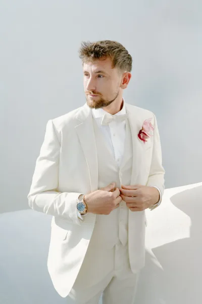 Groom portrait of a groom getting ready, adjusting his bow tie as sunlight washes a white wall backdrop, highlighting his calm, polished look.
