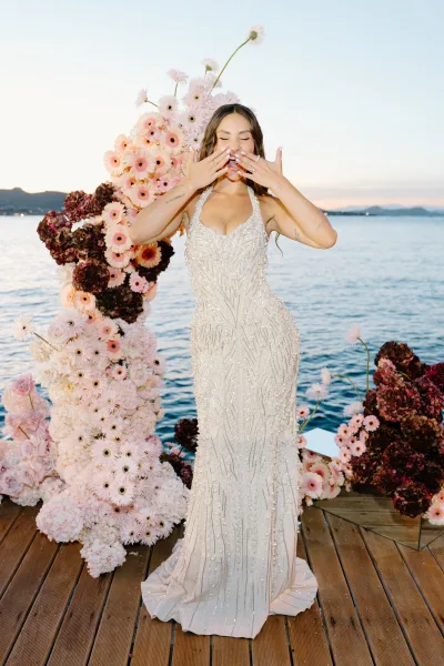 Bridal portrait of a bride blowing a kiss on a wood dock over calm water, backed by mountains and open sky, her long wavy bridal hair catching light.