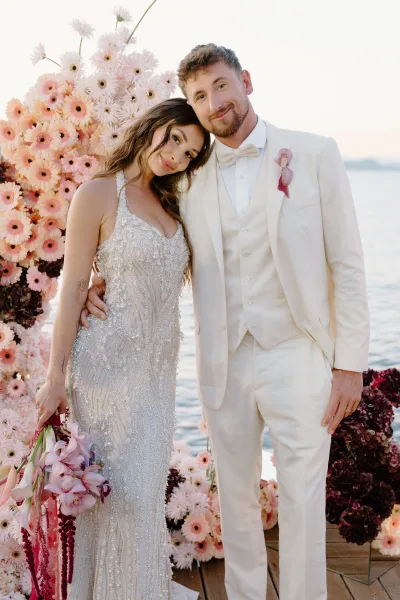 Couple portrait of a bride leaning on groom on a wooden dock by the water, framed by sky, as she holds an orchid bouquet.