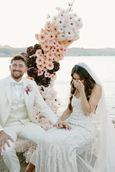 Ceremony moment during wedding vows as the bride and groom stand on a wooden lakeside deck, with a soft bridal veil and distant hills beyond.