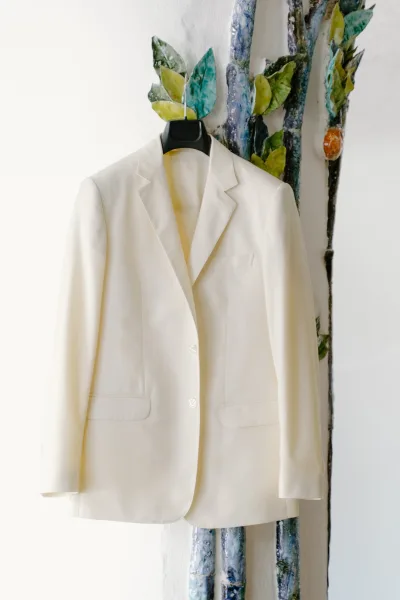 Wedding suit jacket, an ivory groom jacket with a notch lapel, hangs against a white wall adorned with ceramic leaves and a sculptural tree.