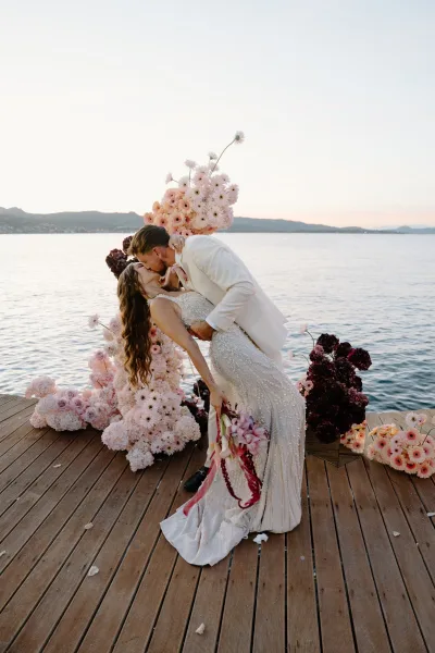 Wedding kiss portrait as the bride and groom dip on a wooden dock by a mountain lake at sunset, the bride holding a cascading orchid bridal bouquet.