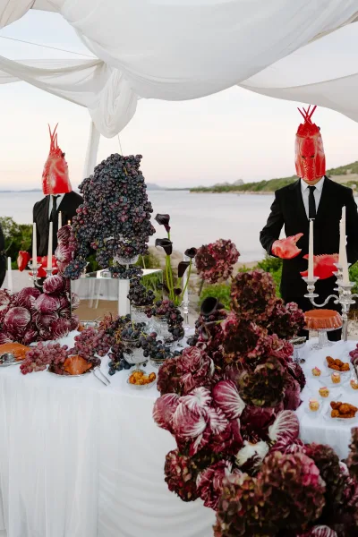 Grazing table and a fruit display table styled with candelabras, set beside the ocean shoreline under open sky and greenery.