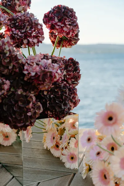 Floral centerpiece in a mirrored vase catching light, set on a wooden deck by the water with open sky and distant hills.