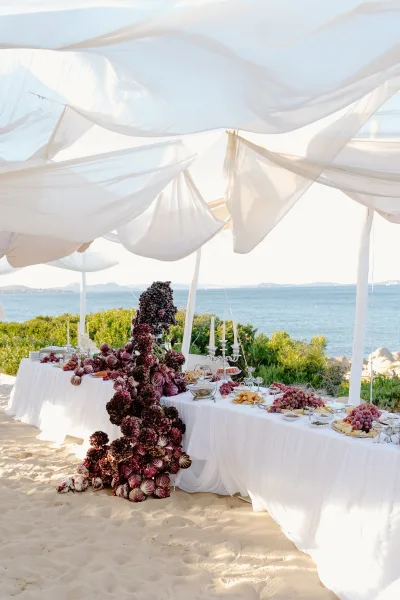 Wedding grazing table at a beach reception buffet, showcased under a draped fabric canopy on sandy shore with ocean and rocks beyond.