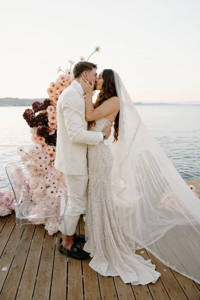 Wedding kiss portrait as they share their ceremony first kiss on a wooden dock, the bride’s veil trailing as water and mountains shimmer behind.