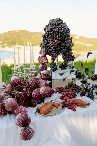 Wedding food display inspiring grazing table ideas, accented by taper candles, set against a beach and ocean backdrop with hills and greenery.