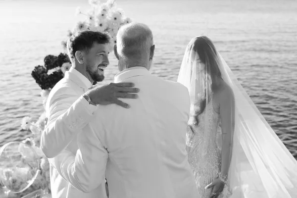 Ceremony moment as the father gives the bride away; the groom hugs him by the water, the bride under a long veil.