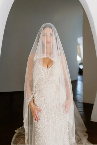 Bridal portrait of a bride with veil in a beaded wedding dress, standing under a white archway in a bright indoor hallway.