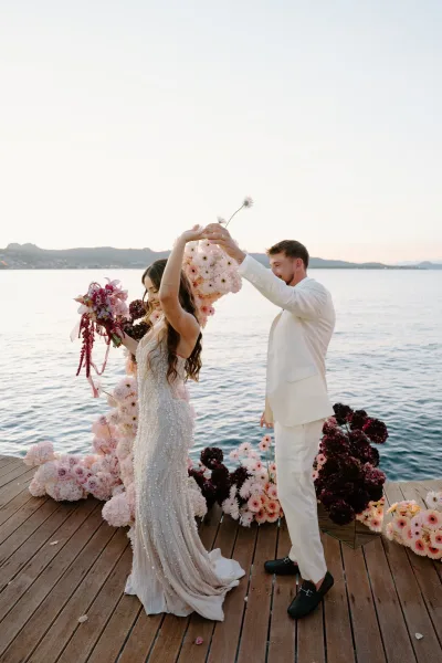 Couple portrait of a bride and groom dancing on a wood deck by the water, with a distant shoreline behind, she holds a pink bridal bouquet.