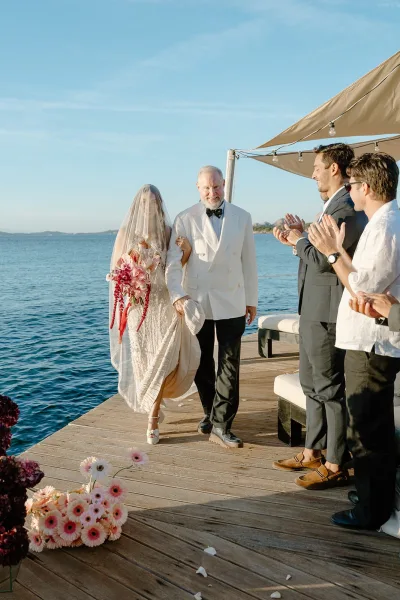 Wedding processional as the bride walks down the aisle holding a cascading orchid bouquet on a wooden dock by the ocean under blue sky.