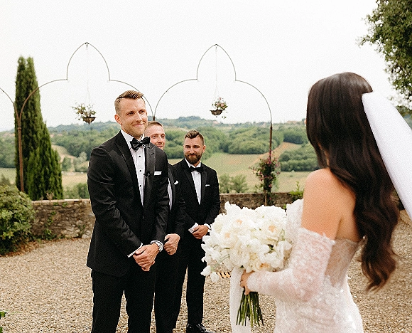 First look moment as bride in strapless lace dress and veil approaches groom in black tuxedo on a terrace by stone wall and hills