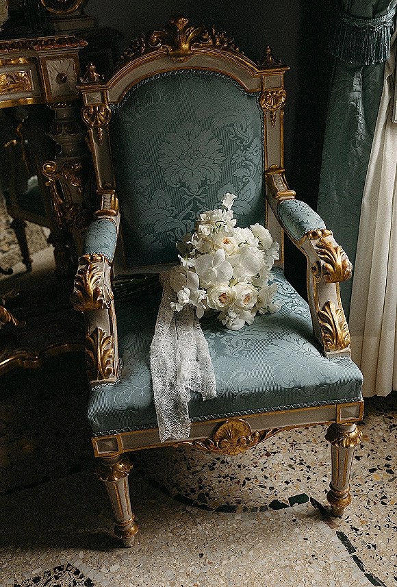 Bridal bouquet, white wedding bouquet with cascading orchids and lace ribbon draped over an upholstered armchair near ornate furnishings