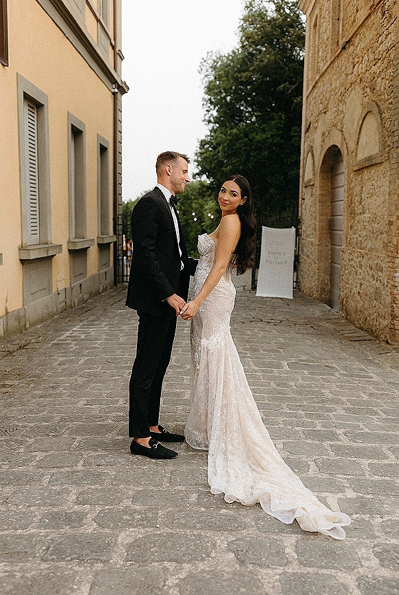 Couple portrait of bride and groom holding hands, bride looking over shoulder with lace train on a cobblestone street by stone buildings
