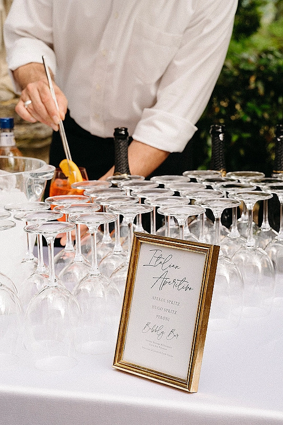 Wedding bar setup with signature cocktail menu sign in a gold frame, coupe glasses and liquor bottles arranged beside greenery outdoors