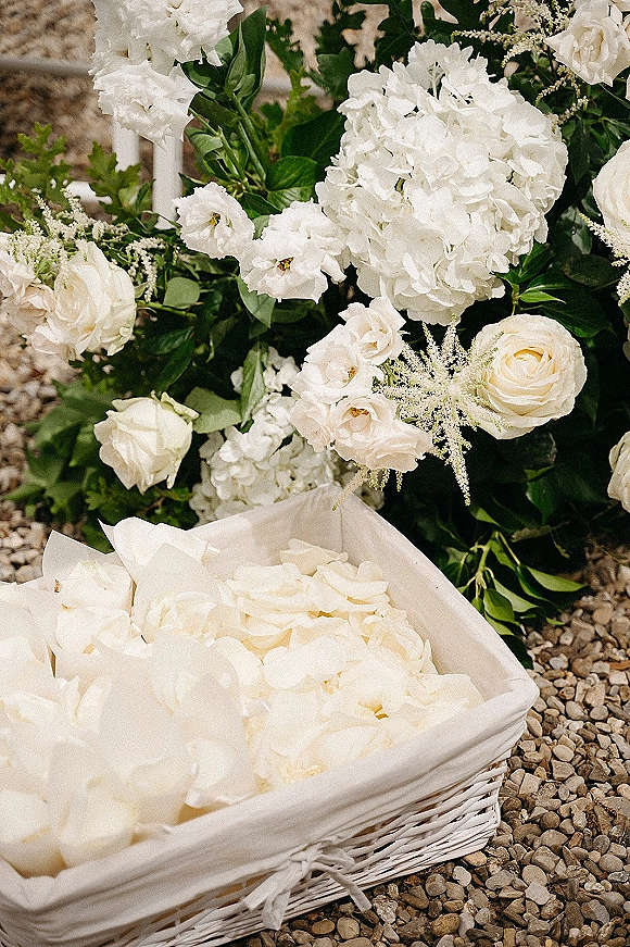 Flower girl basket filled with rose petals and white roses, lined with fabric and tied with a white ribbon, resting on gravel ground