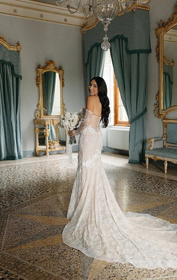 Bridal portrait of bride holding bouquet in an off the shoulder lace wedding dress with long train beneath a chandelier in ornate room