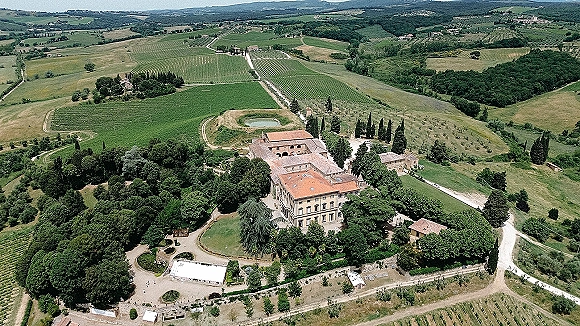 Wedding venue aerial view of a historic villa with terracotta roof, cypress-lined driveway, courtyard, pond, vineyard rows, and tent on rolling hills