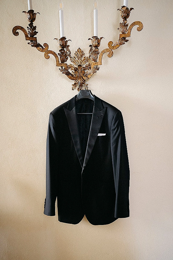 Wedding tuxedo black tuxedo jacket on a hanger with satin lapels and a white pocket square beside taper candles on a beige wall
