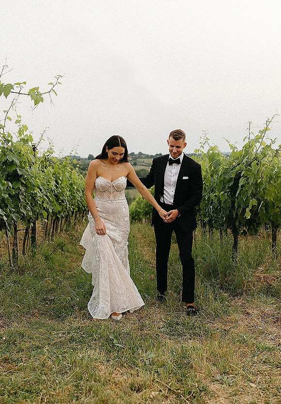 Couple portrait of bride in strapless lace gown and groom in black tux holding hands on a vineyard path under an overcast sky
