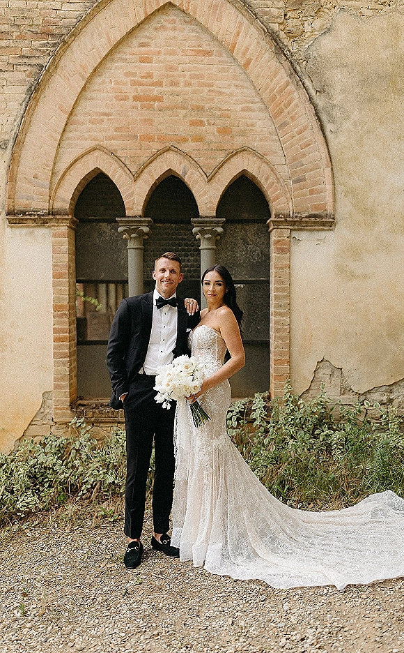 Couple portrait of bride and groom posing under a stone arch, she holds a white bouquet in a lace gown with long veil, he wears a black tuxedo