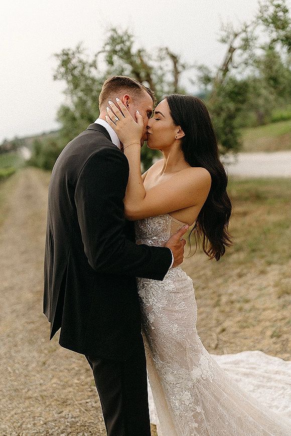 Wedding kiss as bride in a lace mermaid dress cups groom’s face in a black tuxedo on an outdoor path with trees and hillside under overcast sky