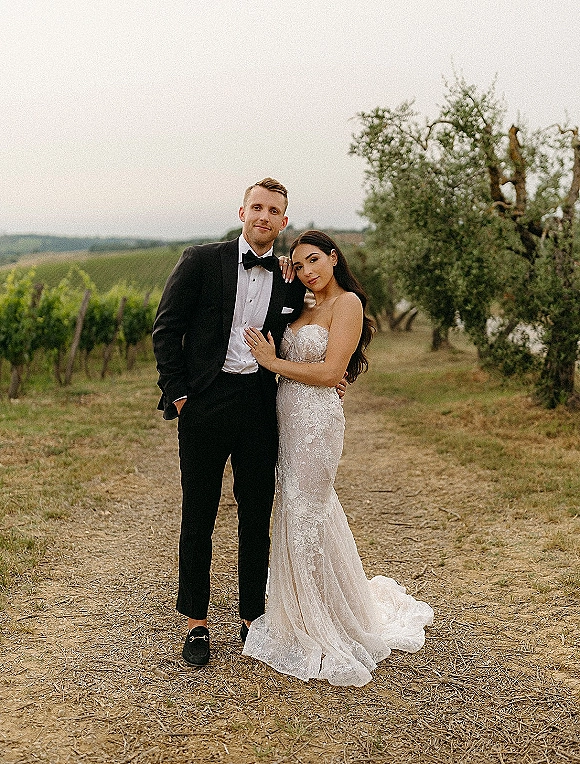 Couple portrait of bride and groom pose on a dirt path between vineyard rows, bride in strapless lace dress resting hand on tuxedoed groom