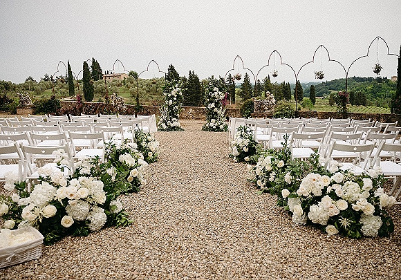Outdoor ceremony setup with wedding aisle flowers lining a gravel aisle, white folding chairs, floral pillars, and stone statues by a stone wall