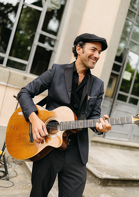 Wedding musician wedding guitarist playing an acoustic guitar in a black suit and flat cap on stone steps outside a windowed building facade
