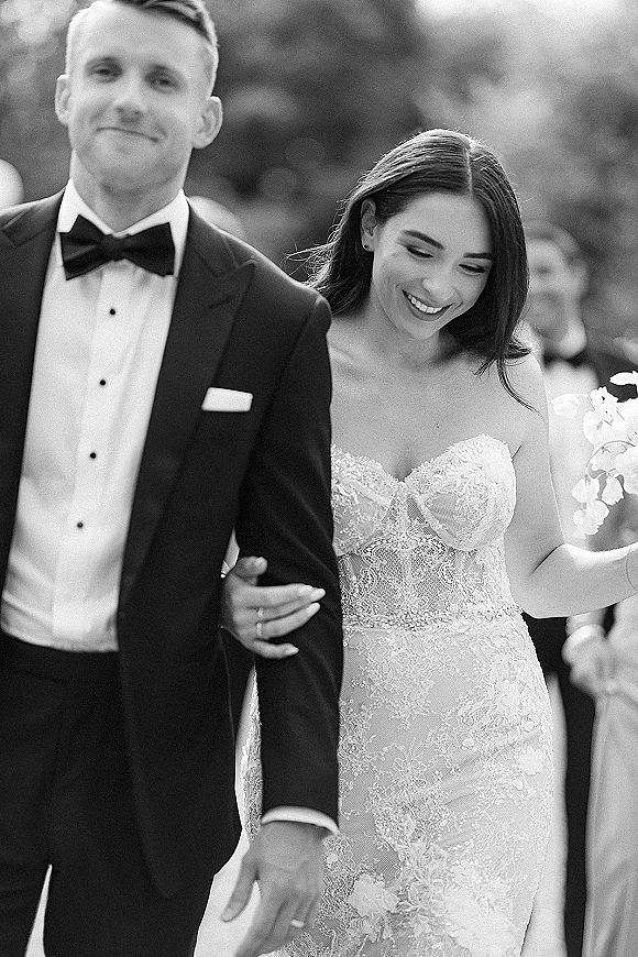 Wedding couple portrait in black and white as bride in a strapless lace gown holds groom’s arm, bouquet in hand, guests blurred in greenery