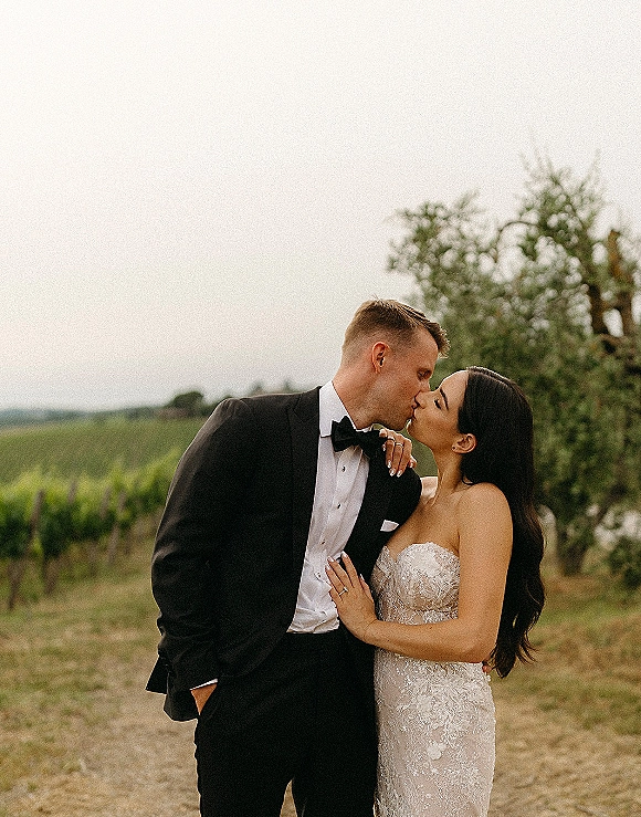 Wedding kiss portrait of bride and groom kissing in a vineyard, her strapless lace dress and his black tuxedo with bow tie under overcast sky