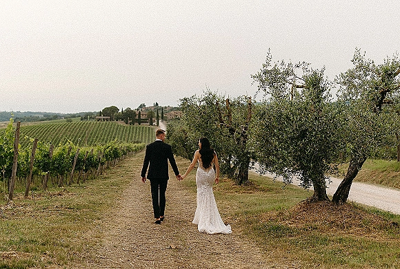 Couple portrait of bride and groom walking away holding hands, her lace wedding dress train and his tuxedo on a vineyard dirt path