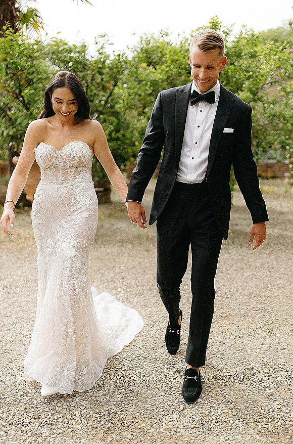 Couple portrait of bride and groom walking hand in hand, her strapless lace dress train flowing beside his black bow tie on a garden path