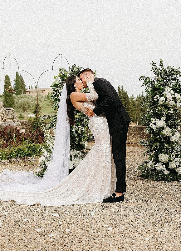 Wedding kiss under a greenery wedding arch, bride in lace mermaid dress and veil, groom in black suit by stone wall and villa hillside
