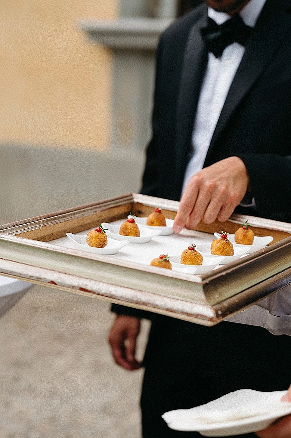 Wedding passed appetizers on white tasting spoons held by a tuxedo server with bow tie on a tray in an outdoor courtyard by stone walls