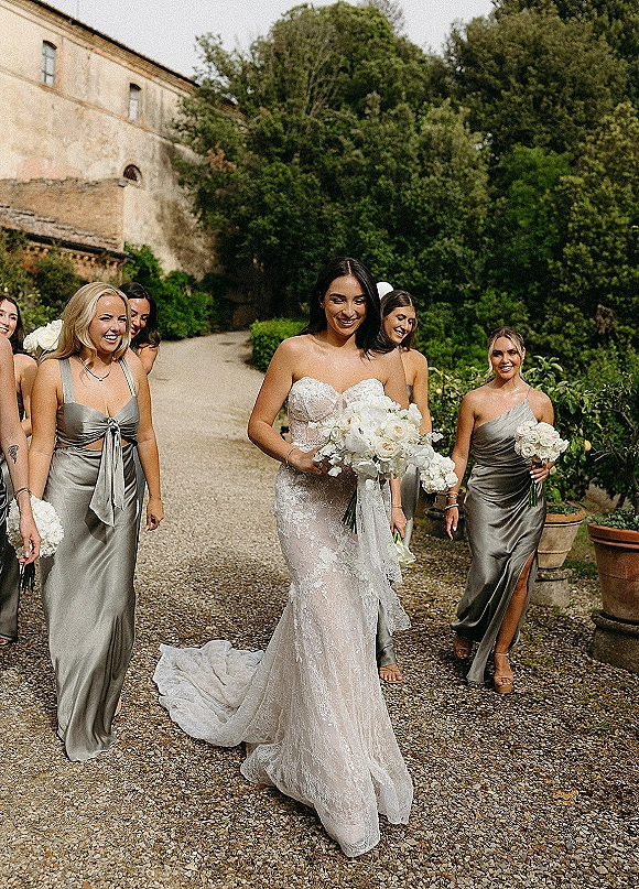 Bride with bridesmaids walking together, holding white rose bouquets in silver satin dresses on a gravel path beside greenery and stone building