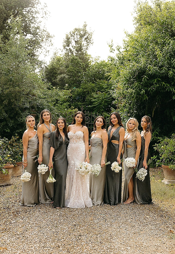 Bridesmaids group photo with bride, satin mismatched dresses and white bouquets, posed on a gravel garden path by a wrought iron gate