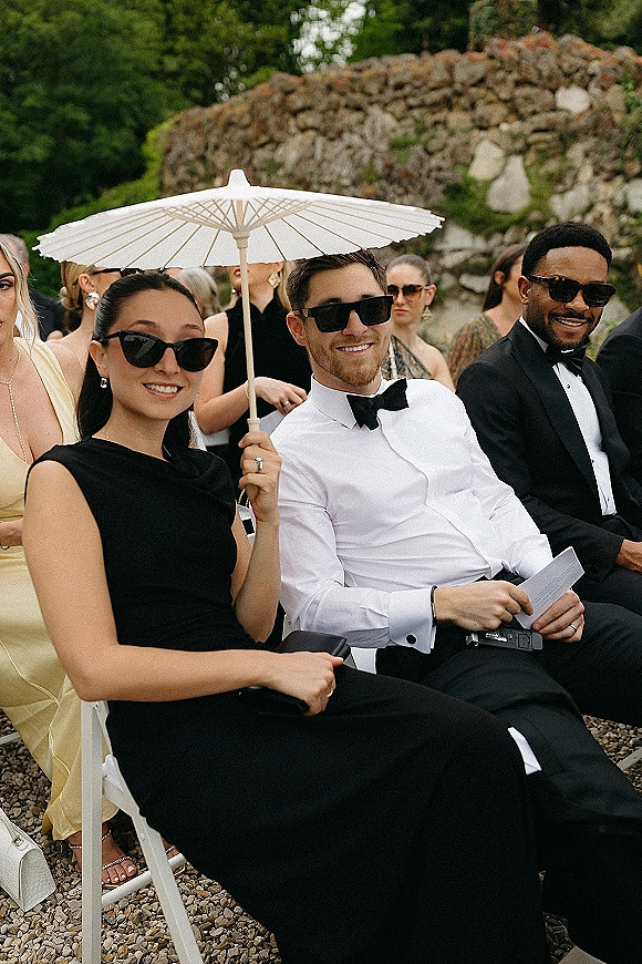 Wedding guests in black tie attire, one holding a white paper parasol, seated on white folding chairs at an outdoor ceremony by a stone wall