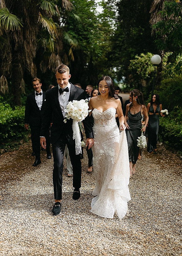 Wedding recessional with bride and groom walking hand in hand, bride holding a bouquet on a tree-lined gravel garden path with palms