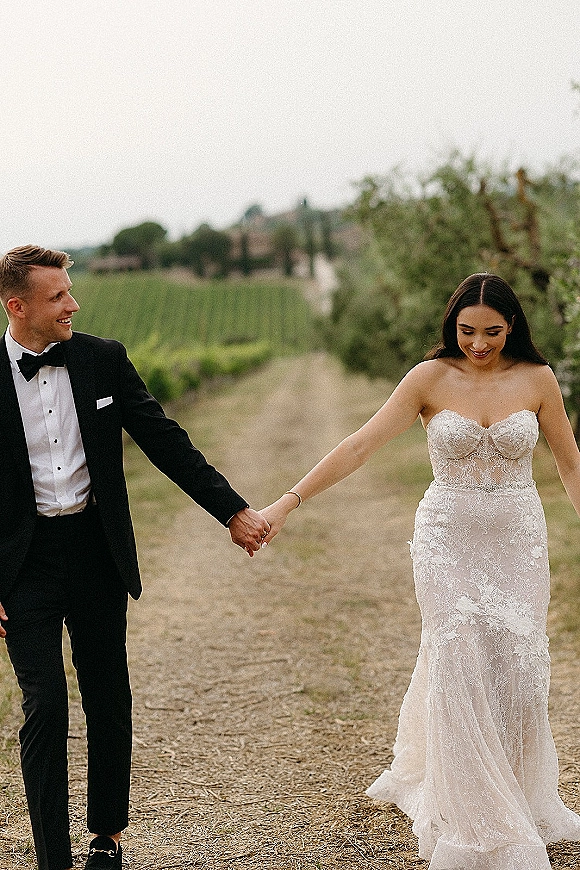 Couple portrait of bride and groom holding hands, walking a vineyard dirt road under an overcast sky, her lace strapless dress and his tuxedo bow tie