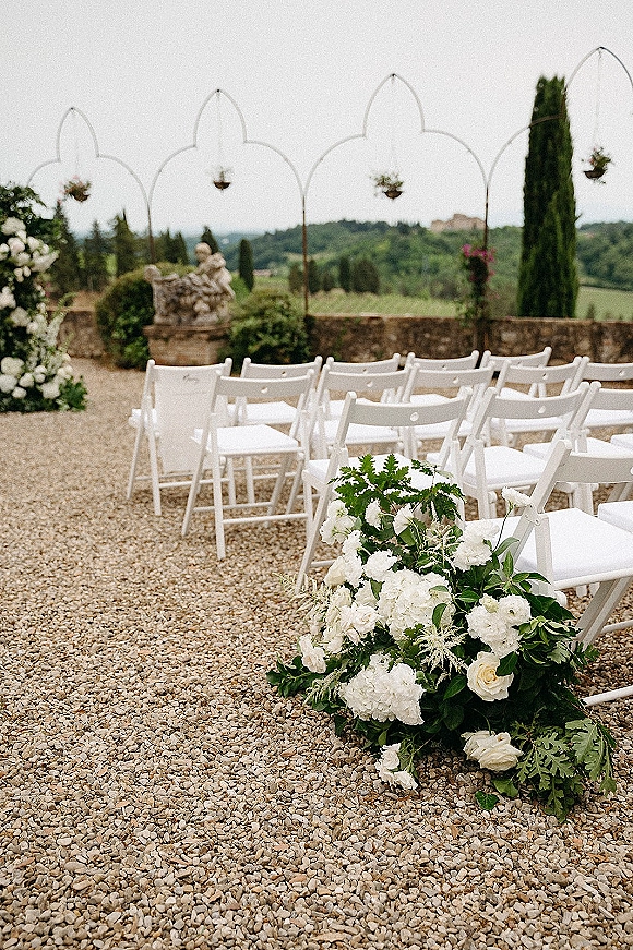 Ceremony setup with white folding chairs lining a gravel aisle, low white and green florals, and arch frames in a stone-walled courtyard
