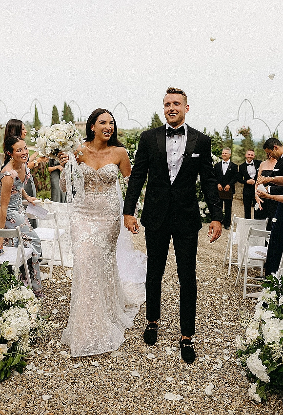 Wedding recessional as bride and groom walk the aisle hand in hand, bouquet and veil trailing past guests, petals, and garden arch frames