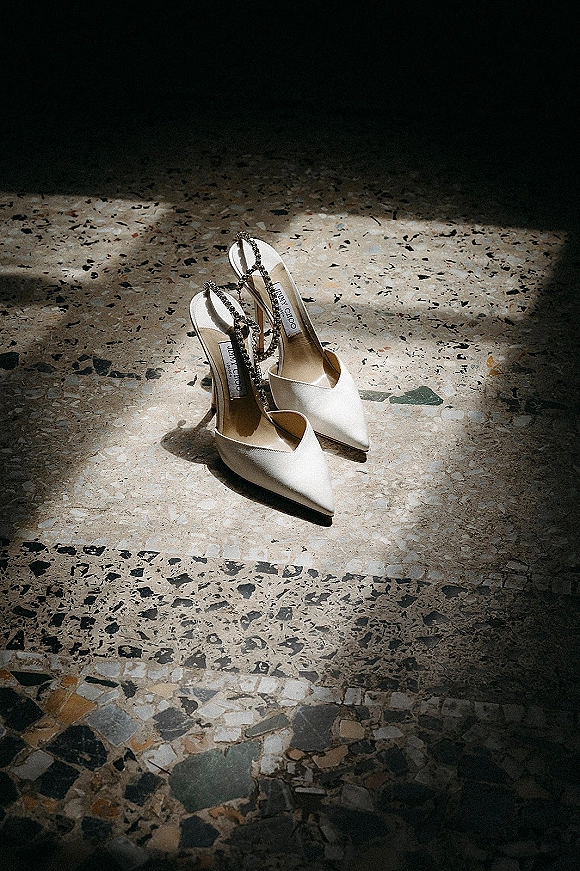 Bridal shoes, ivory wedding heels with crystal ankle straps on a terrazzo floor, highlighted by a dramatic spotlight shadow