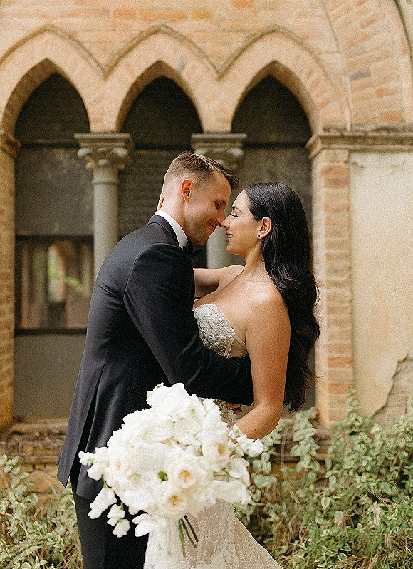 Couple portrait of bride and groom embrace with a forehead touch, holding a white rose bouquet under stone arches in a courtyard