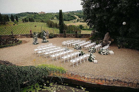Ceremony setup for an outdoor wedding ceremony with white folding chairs, floral arch, and greenery in a gravel courtyard overlooking vineyard hills